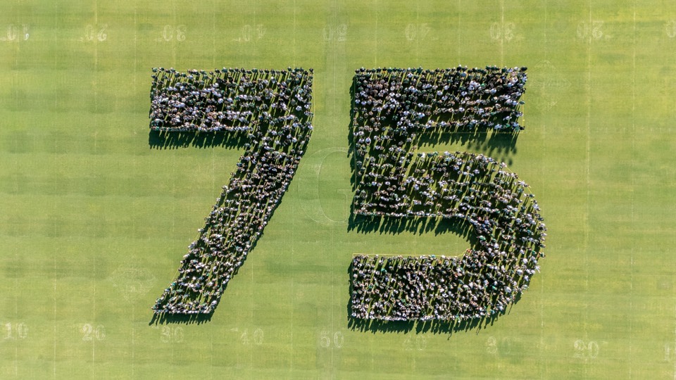 Aerial of hundreds of people forming the number seventy-five on a green field